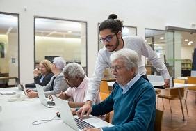 Young male teacher with bun and beard leaning over older male student to point out something on a laptop